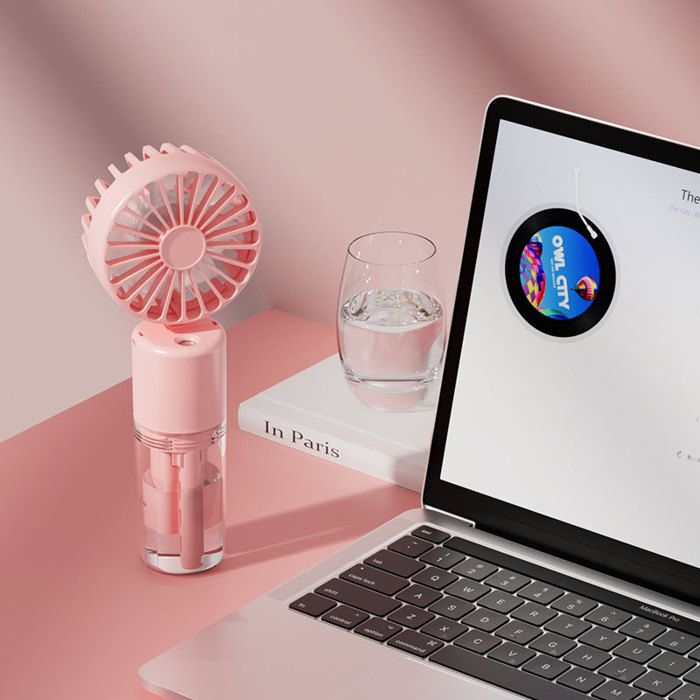 Le même ventilateur rose pâle est posé sur une surface rose, à côté dun ordinateur portable ouvert affichant un logo OWL CITY, dun verre deau et dun livre intitulé In Paris. Le fond est rose uni. Le texte In